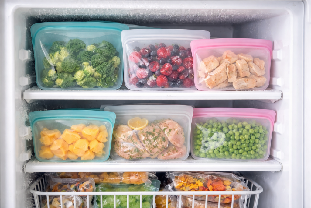 Organized freezer with reusable silicone food storage bags filled with frozen broccoli, berries, chicken, mango, and peas, demonstrating freezer-safe, eco-friendly food storage alternative to plastic bags.