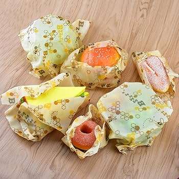 Assortment of foods wrapped in reusable beeswax wraps on a wooden table, including a sandwich, bread slice, apple, donut, and small containers, showing how wraps can be folded around different shapes.
