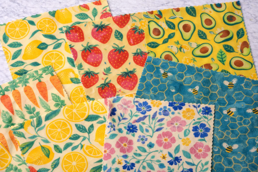 Collection of colorful beeswax wraps laid flat on a surface, featuring patterns of lemons, strawberries, carrots, avocados, honeycomb, and flowers in bright, cheerful designs.